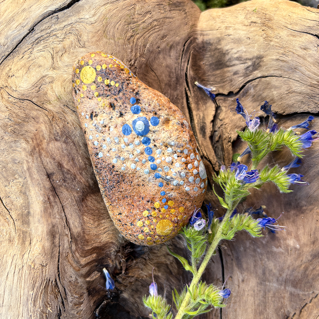 Rückseite von Engel auf Stein mit Acrylfarbe gestaltet auf einer Wurzel umrahmt von blauen Blumen