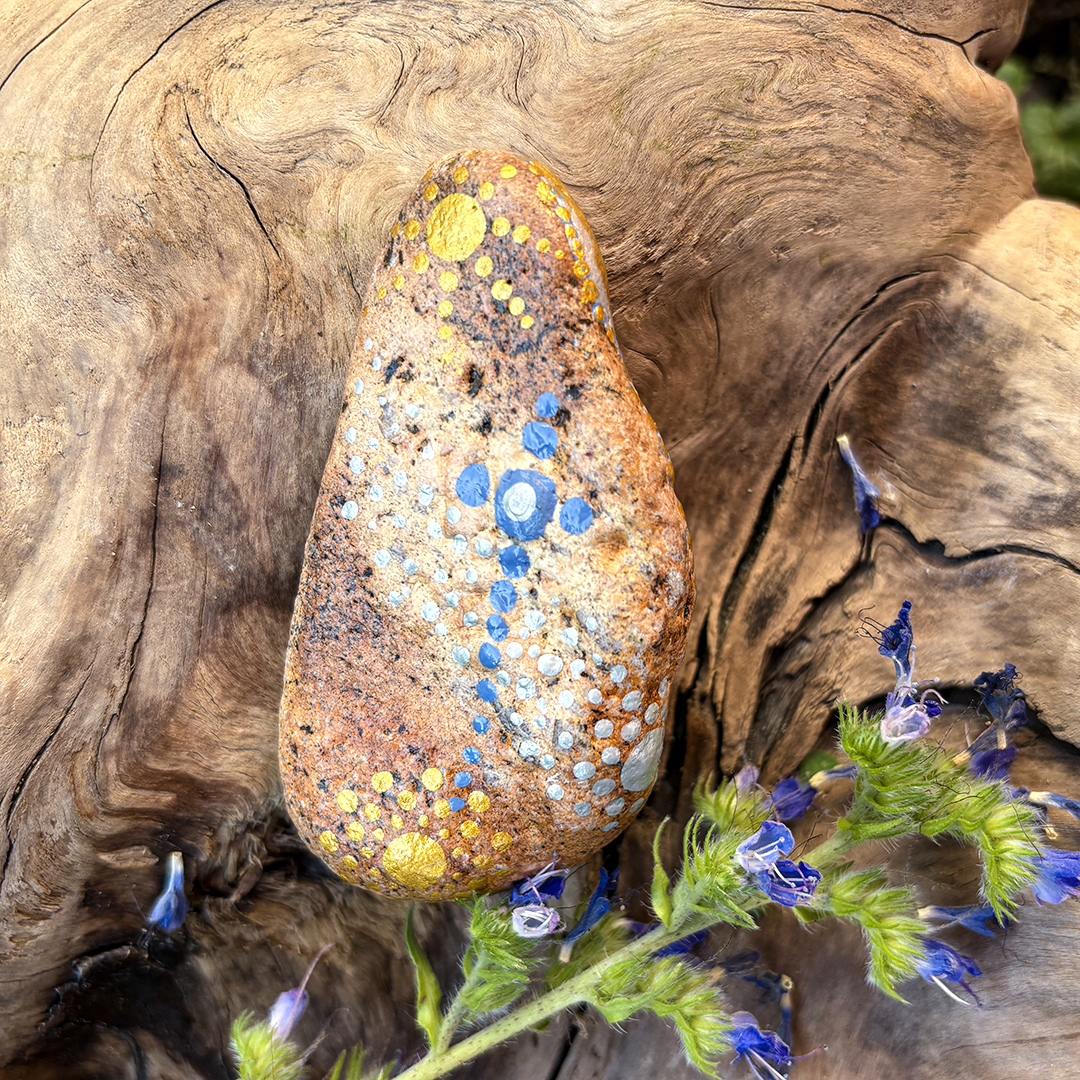 Rückseite eines Engel auf Stein mit Acrylfarbe gestaltet auf einer Wurzel umrahmt von blauen Blumen