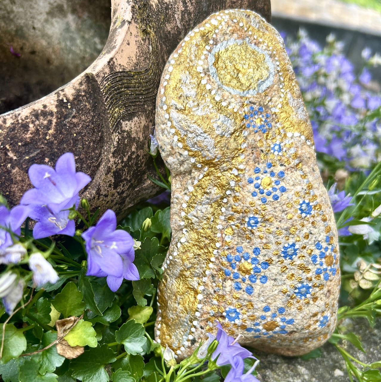 Engel auf Stein mit Acrylfarbe gestaltet vor Tonamphore und blauen Blumen