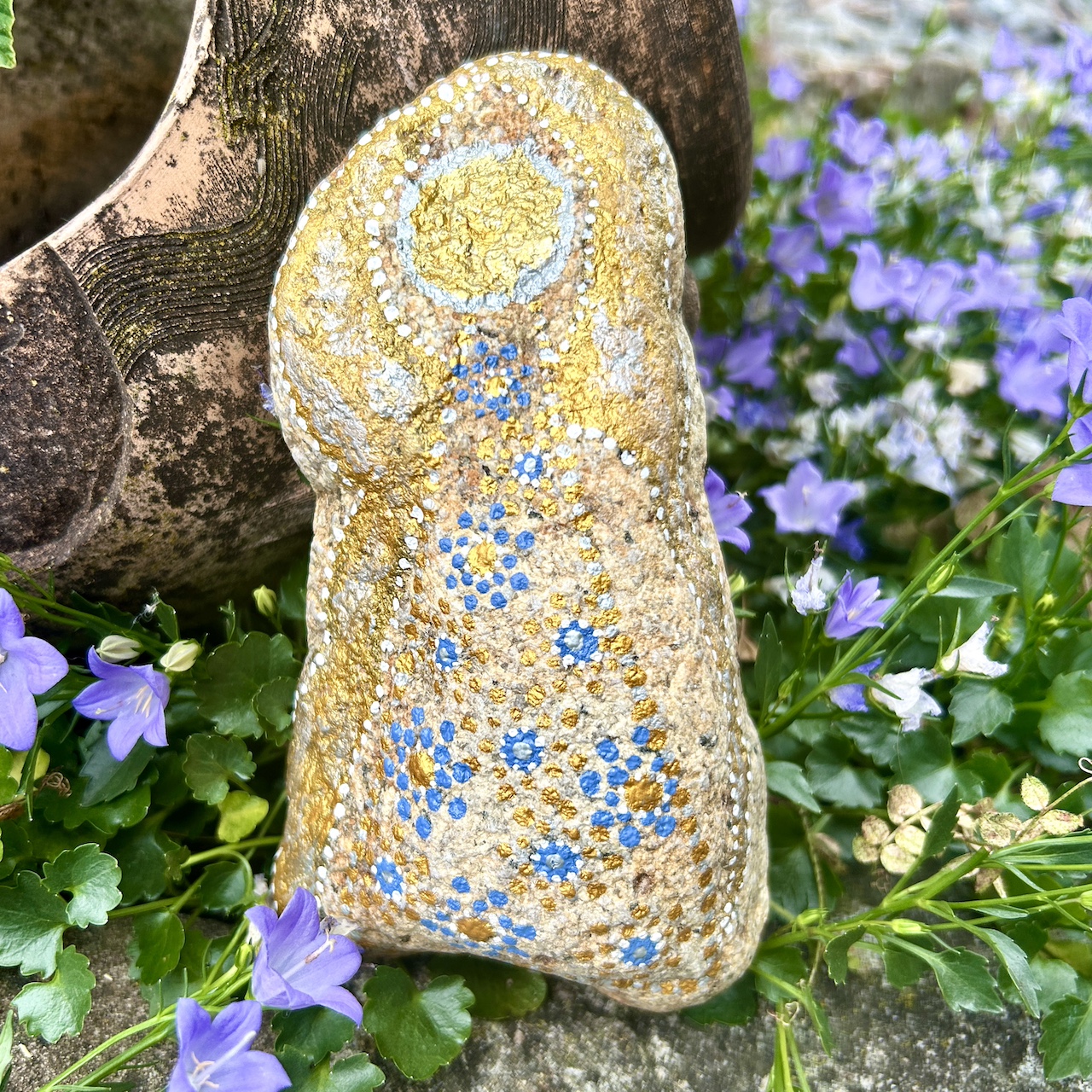Engel auf Stein mit Acrylfarbe gestaltet vor Tonamphore und blauen Blumen