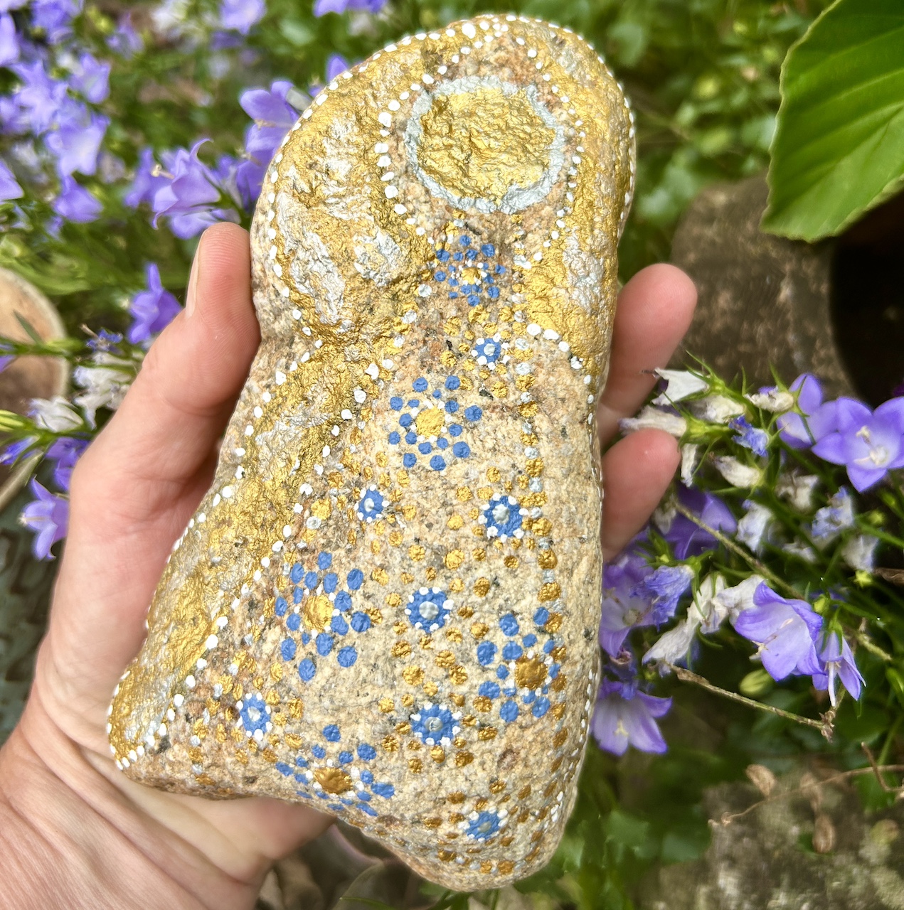Engel auf Stein mit Acrylfarbe gestaltet in der Hand vor blauen Blumen 