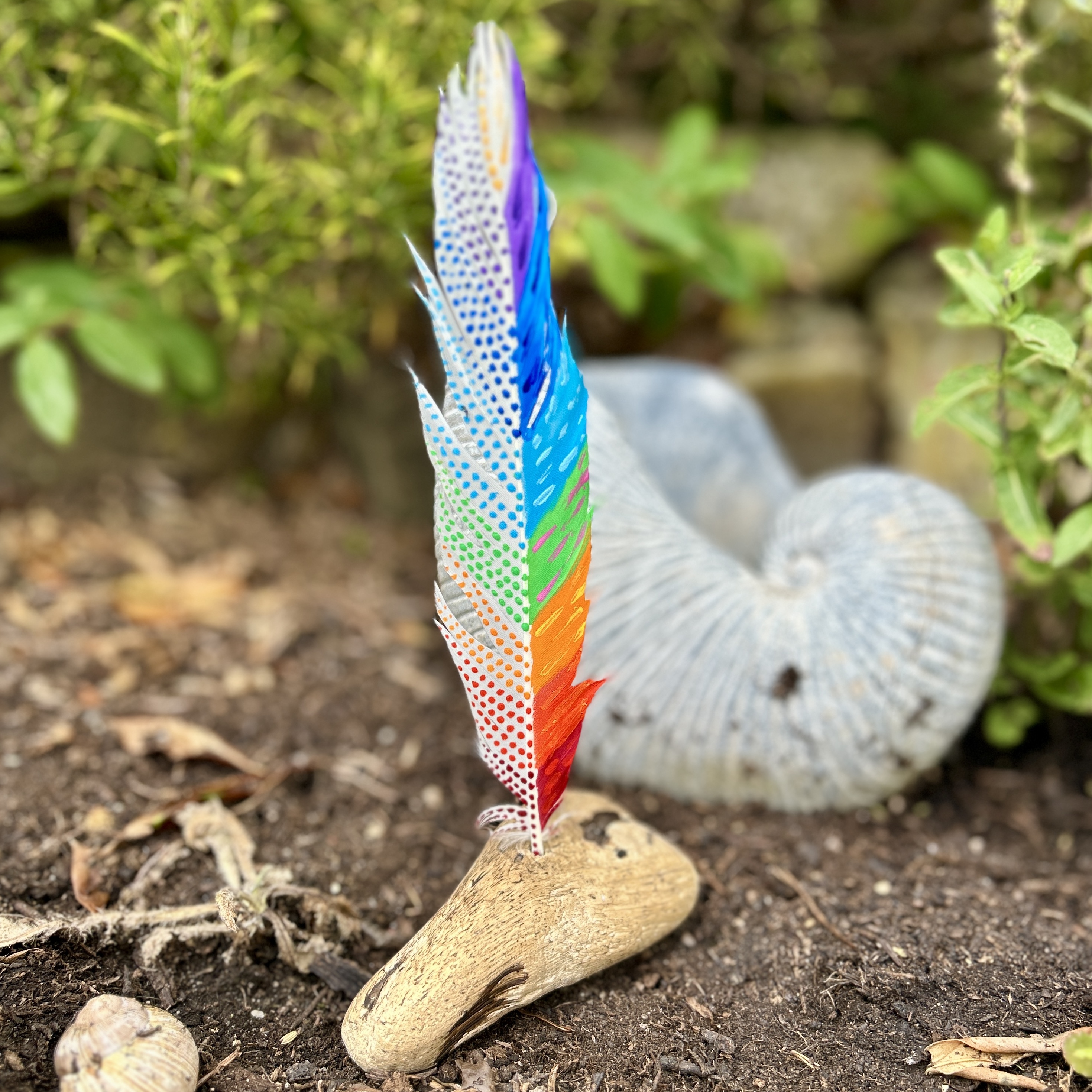 eine bunt in den Farben der Chakren bemalte Feder in einem Holzständer im Garten