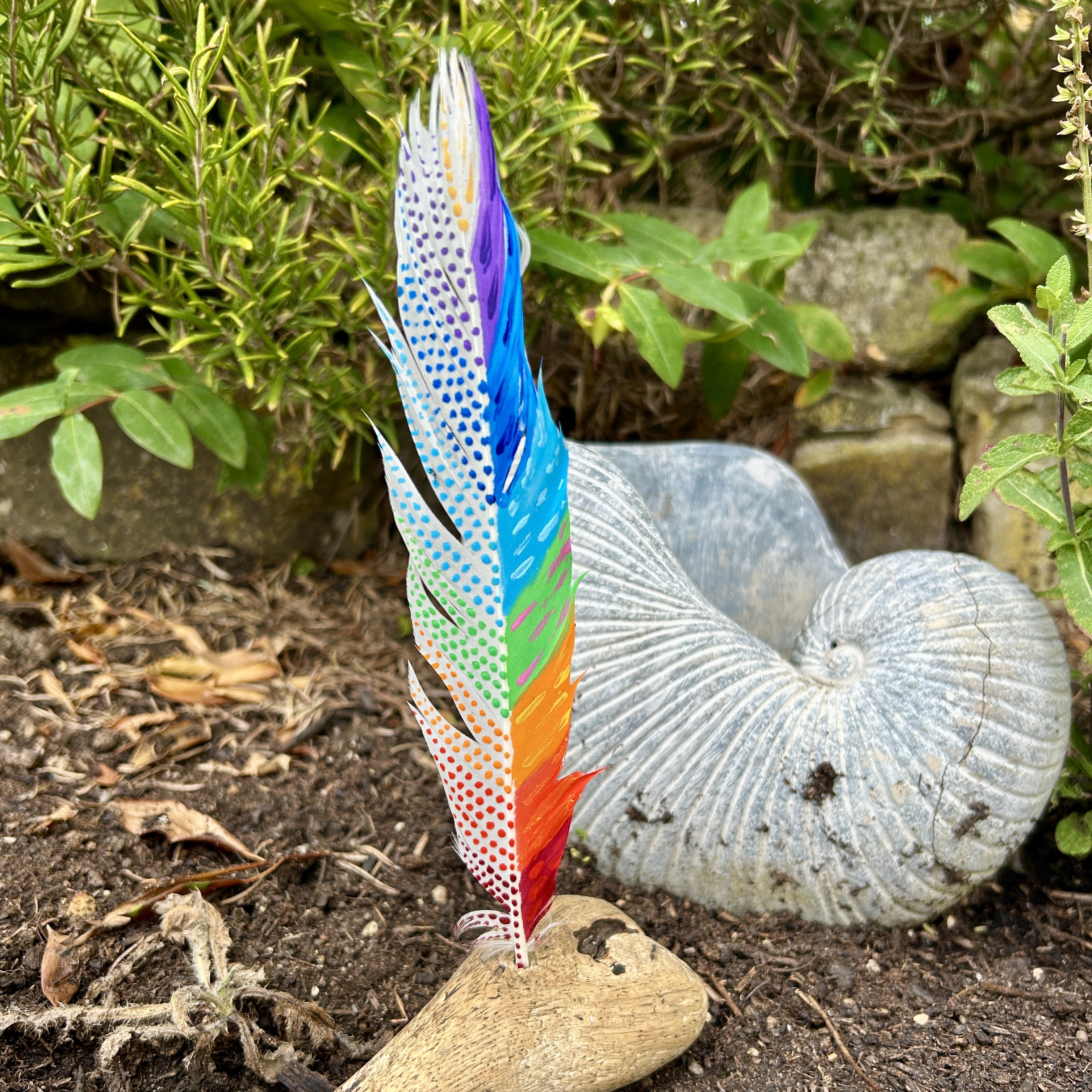 eine bunt in den Farben der Chakren bemalte Feder in einem Holzständer im Garten
