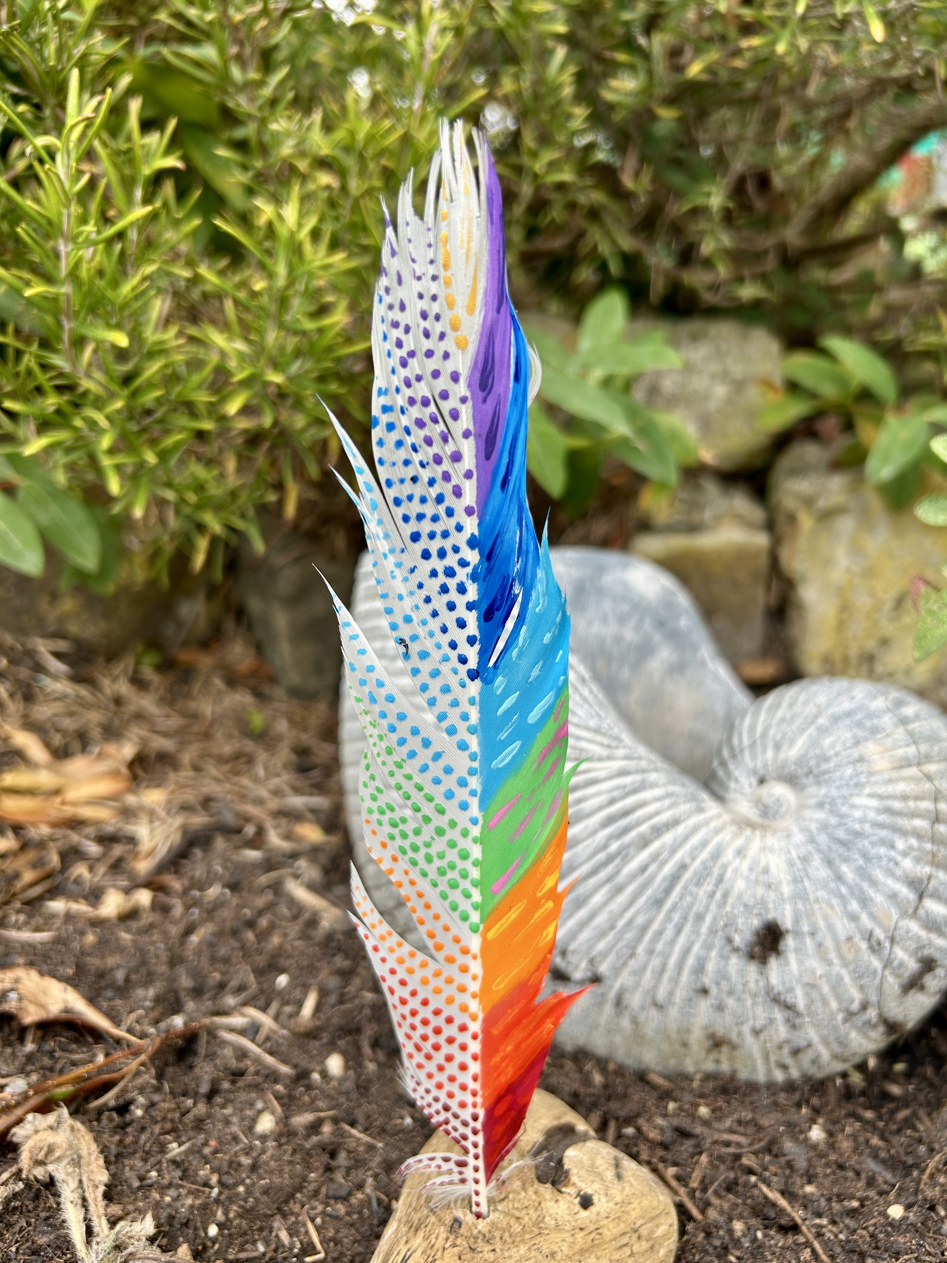 eine bunt in den Farben der Chakren bemalte Feder in einem Holzständer im Garten