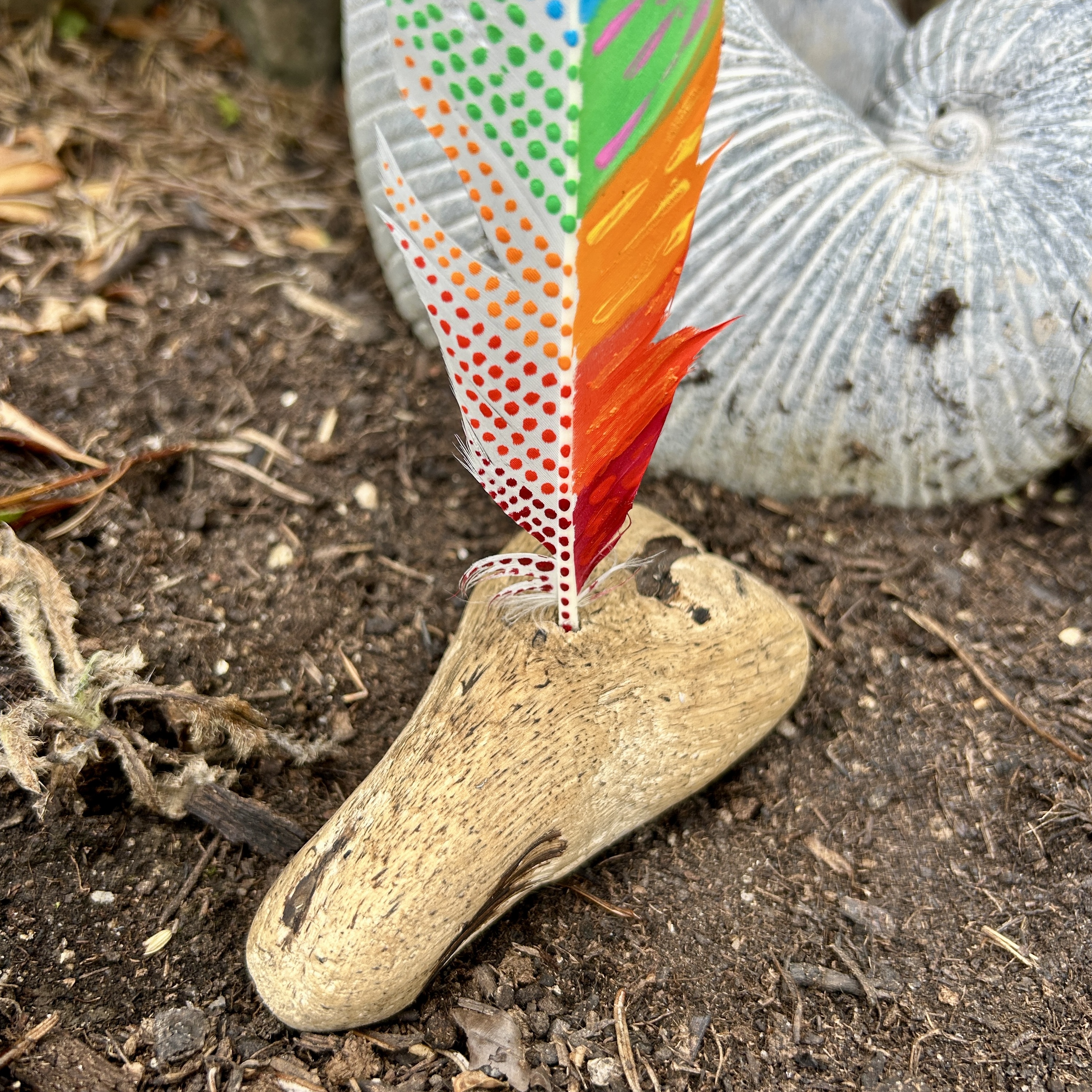eine bunt in den Farben der Chakren bemalte Feder in einem Holzständer im Garten