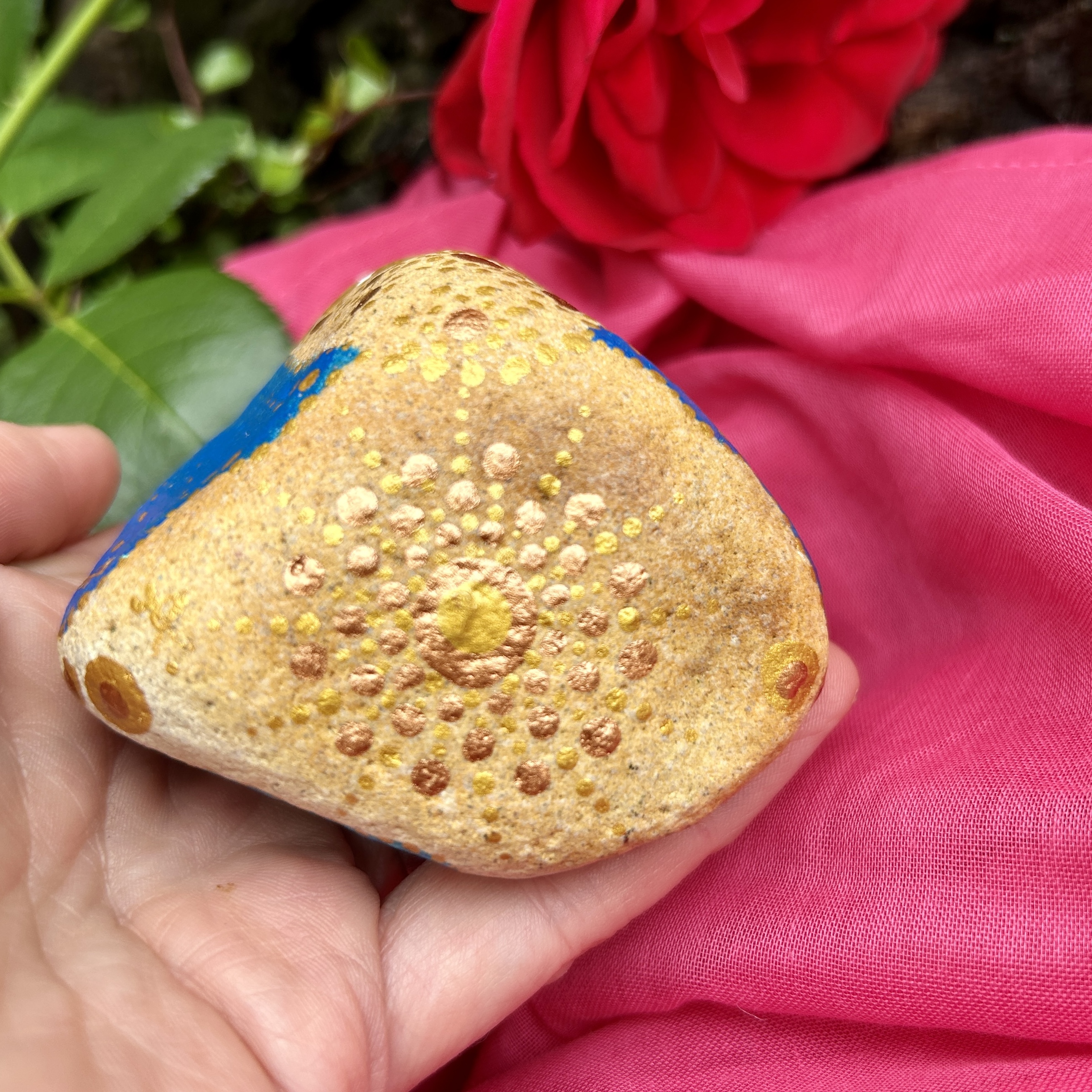 Stein mit Mandala in Gold bemalt auf rosa Tuch vor roter Rose