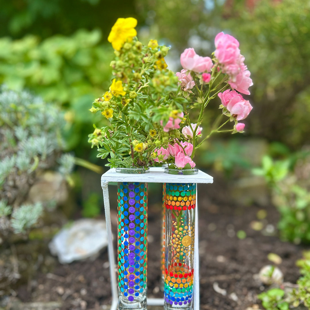 Glasvasen bemalt mit Mandalas in Regenbogenfarben mit rosa und gelben Blumen darin