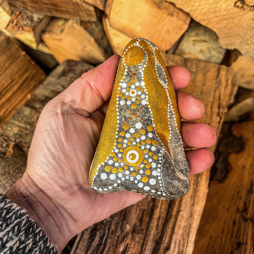 Hand, die einen Stein mit Engel in Gold und Weiß bemalt vor Holzscheiten hält