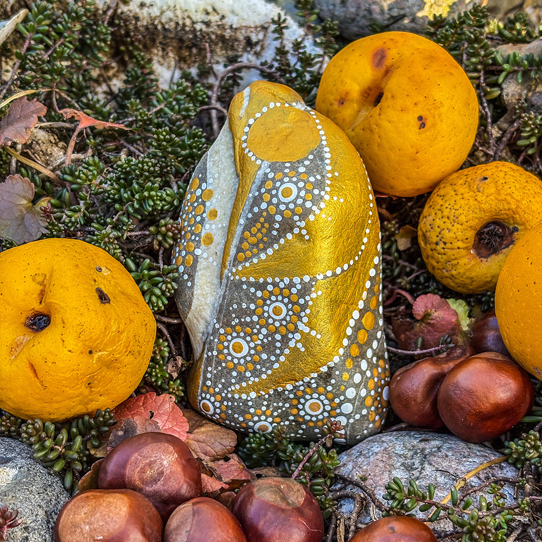 Stein bemalt mit einem Engel in Gold und Weiß zwischen Zierpfirsichen und Kastanien liegend