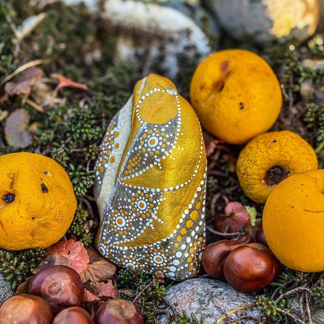 Stein bemalt mit einem Engel in Gold und Weiß zwischen Zierpfirsichen und Kastanien liegend