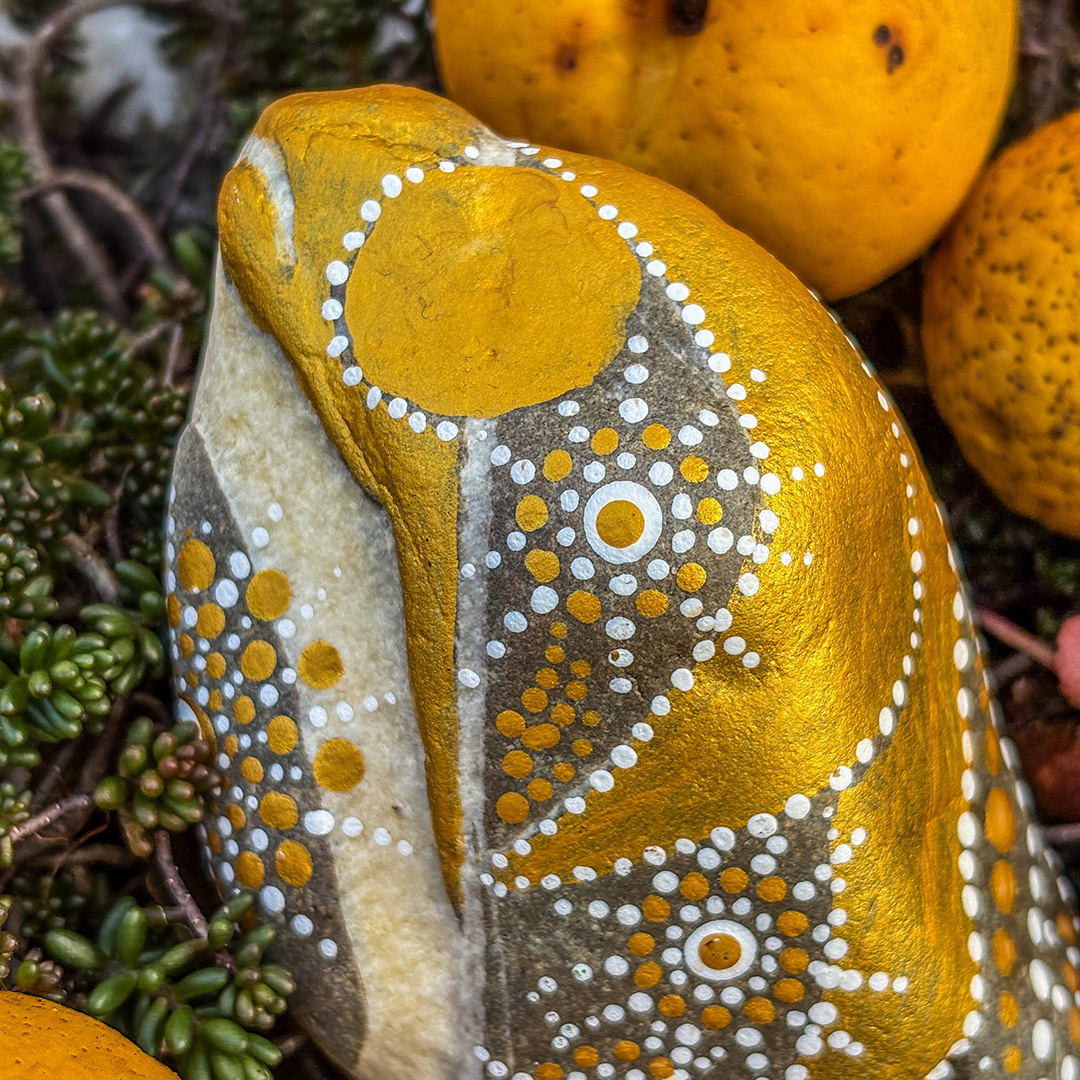 Nahaufnahme von Stein bemalt mit einem Engel in Gold und Weiß neben Zierpfirsichen liegend