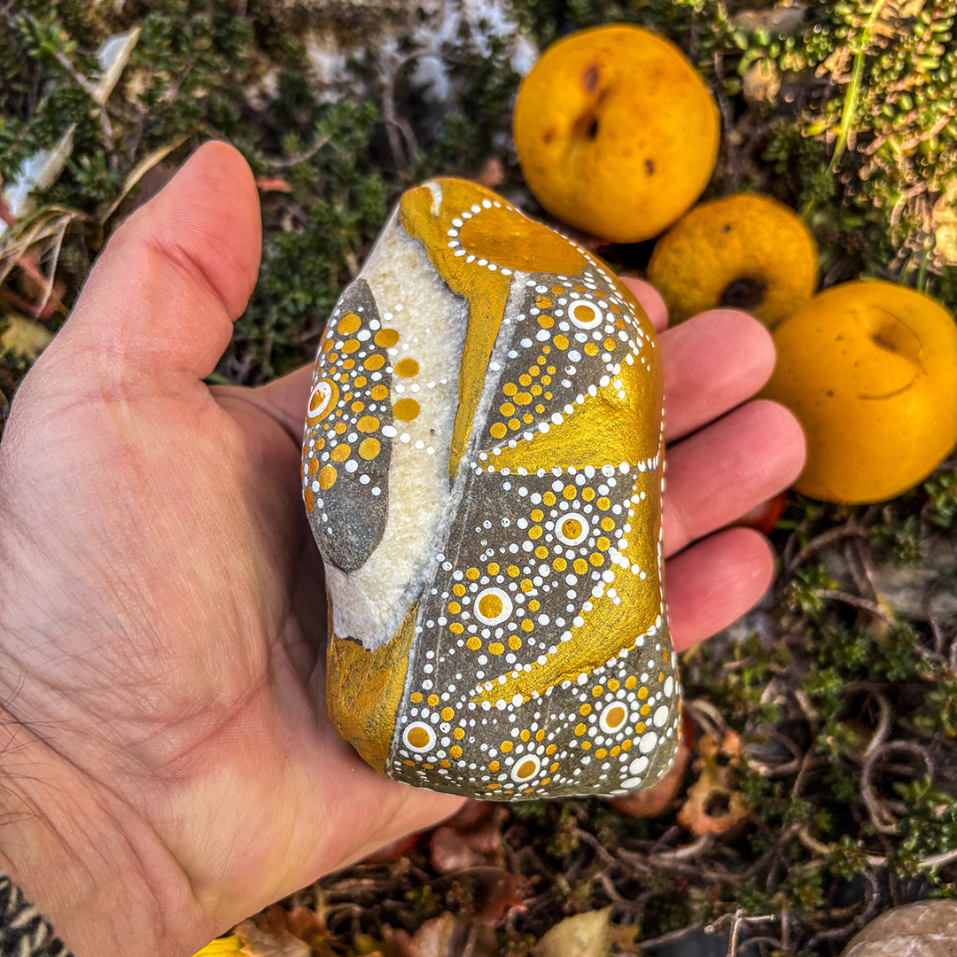 Hand, die einen Stein hält bemalt mit einem Engel in Gold und Weiß vor Zierpfirsichen 