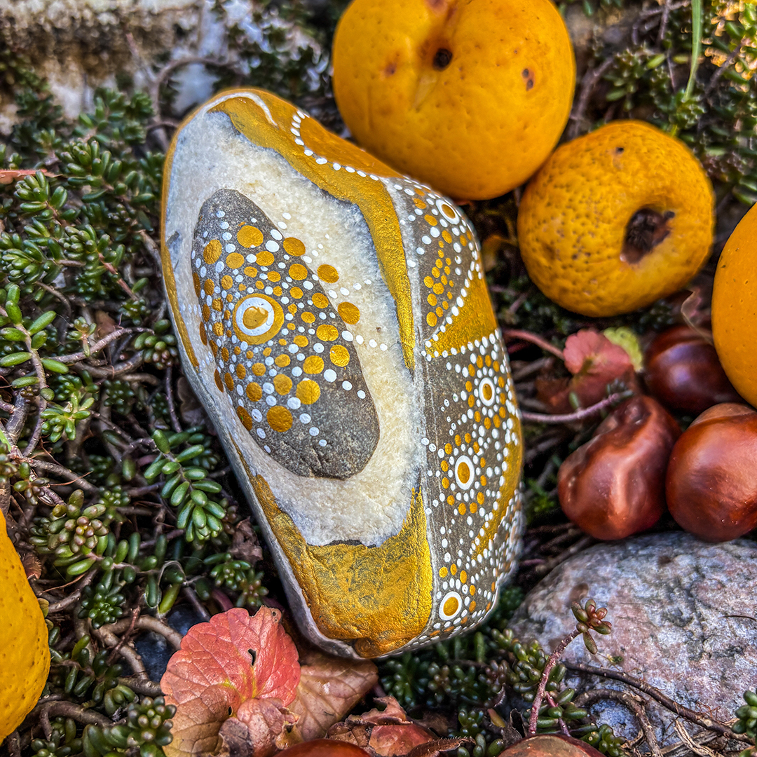 Stein bemalt mit einem Engel in Gold und Weiß zwischen Zierpfirsichen und Kastanien liegend