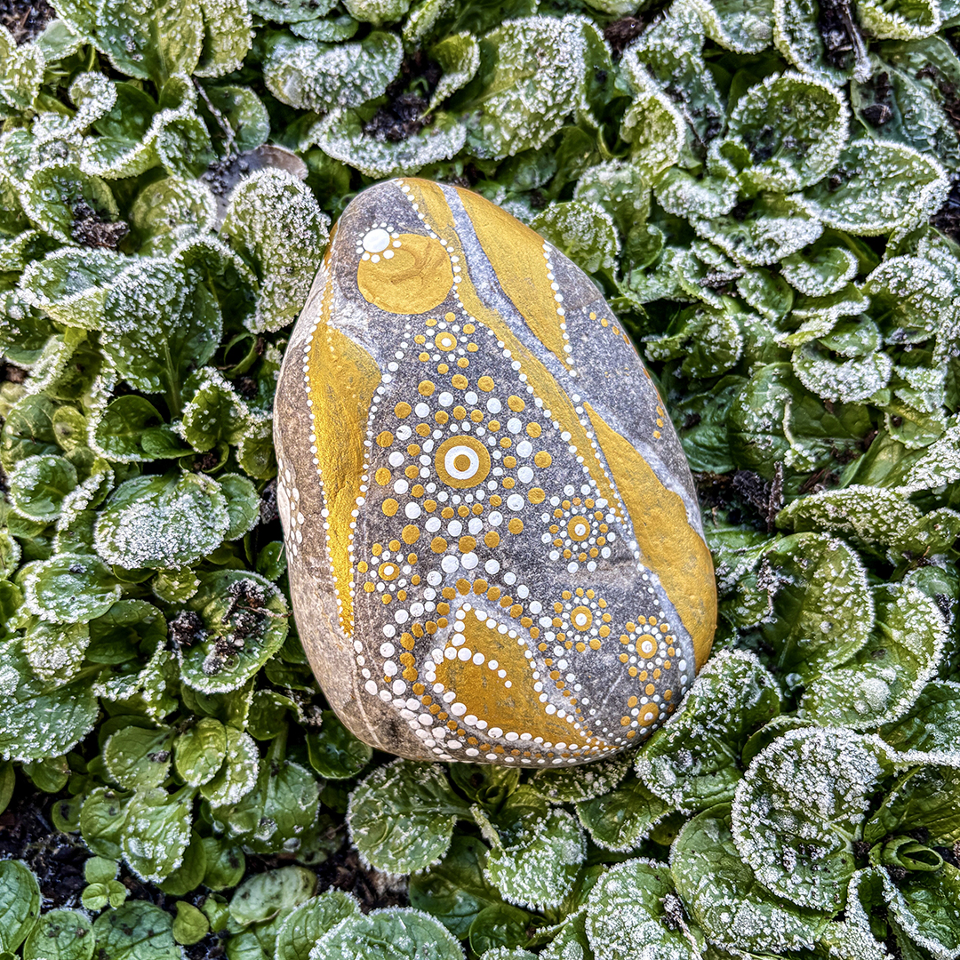 Stein bemalt mit einem Engel in Gold und Weiß auf frostigen Blättern liegend