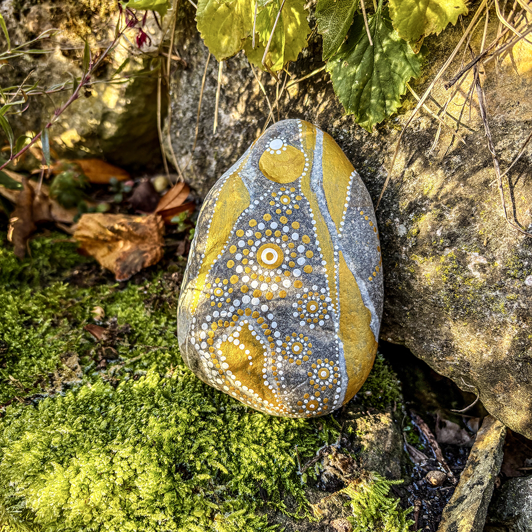 Stein bemalt mit einem Engel in Gold und Weiß auf Moos vor Steinen stehend