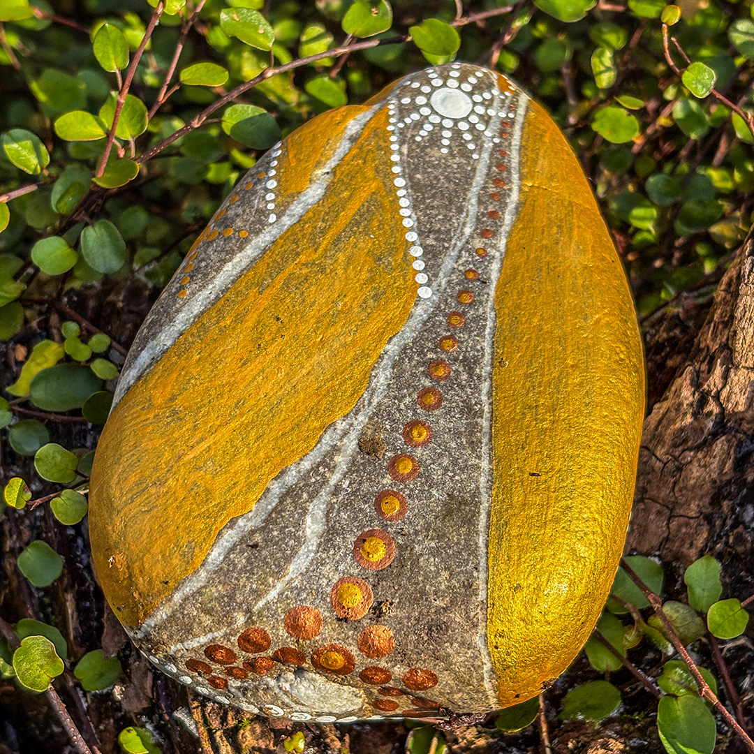 Stein bemalt in Gold und Weiß auf Wurzel zwischen Blättern liegend