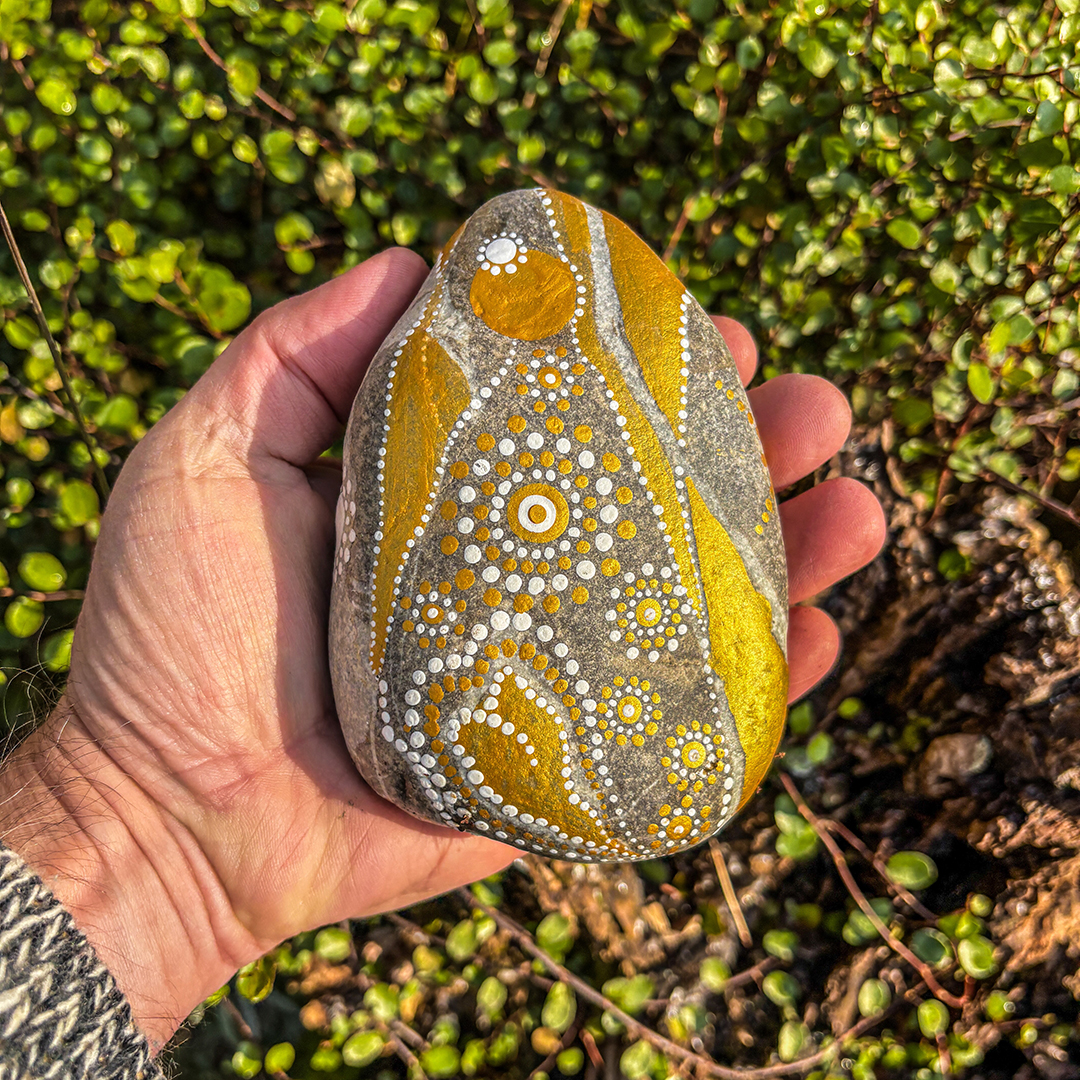 Hand, die einen Stein hält bemalt mit einem Engel in Gold und Weiß vor Blättern