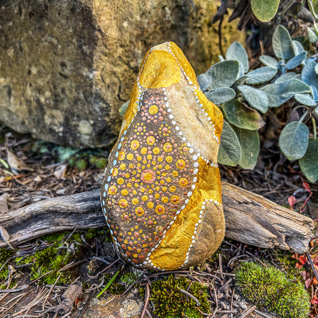 Stein bemalt mit einem Engel in Gold und Weiß vor Steinen, Blättern und Ast