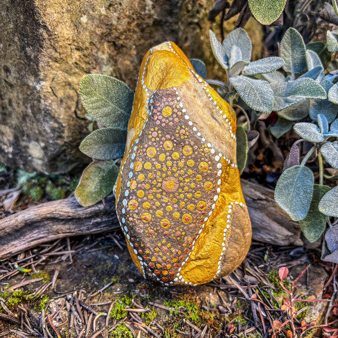 Stein bemalt mit einem Engel in Gold und Weiß vor Steinen, Blättern und Ast