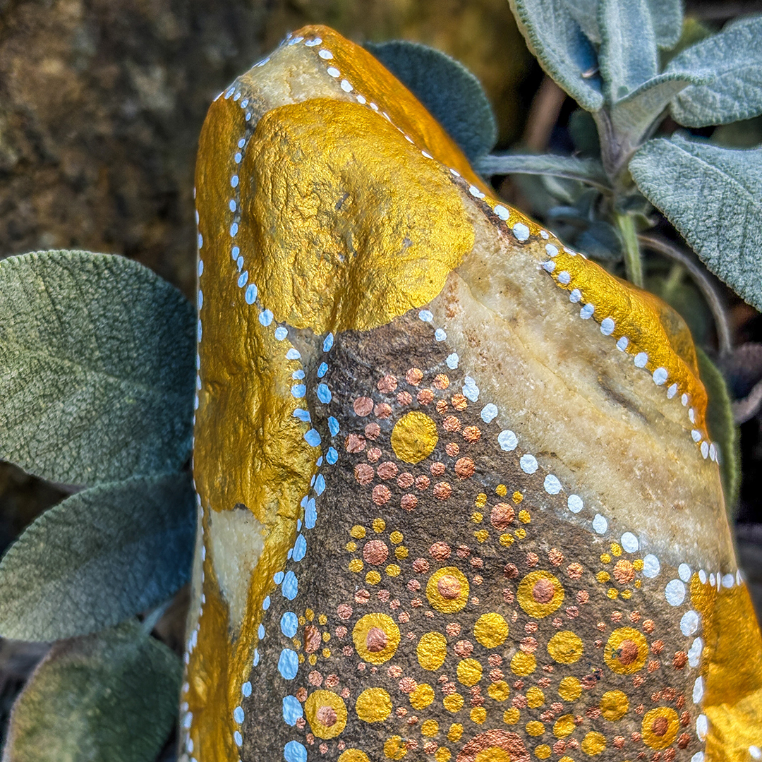 Nahaufnahme eines Steins bemalt mit einem Engel in Gold und Weiß vor Steinen, Blättern und Ast