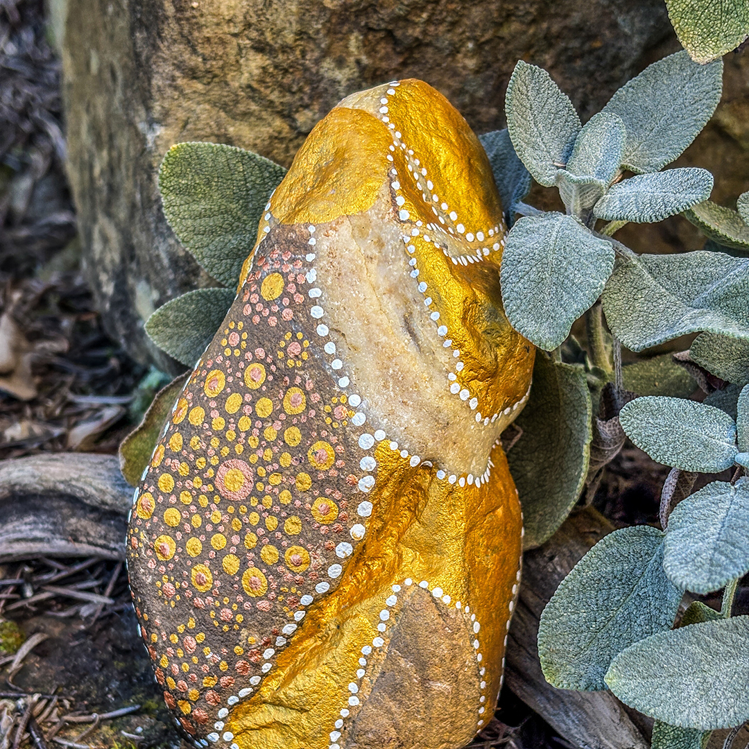 Stein bemalt mit einem Engel in Gold und Weiß vor Steinen, Blättern und Ast
