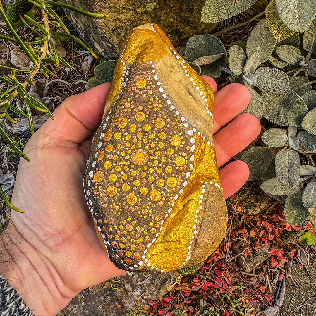Hand hält einen Stein bemalt mit einem Engel in Gold und Weiß vor Steinen, Blättern und Ast