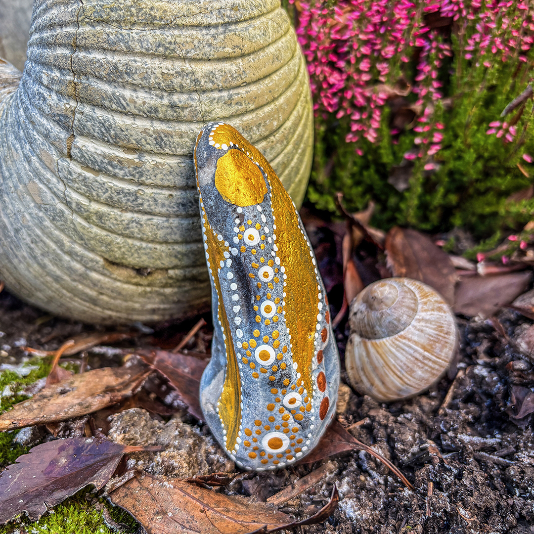 Bemalter Stein mit Engel vor roter Erikapflanze neben Schneckenhaus