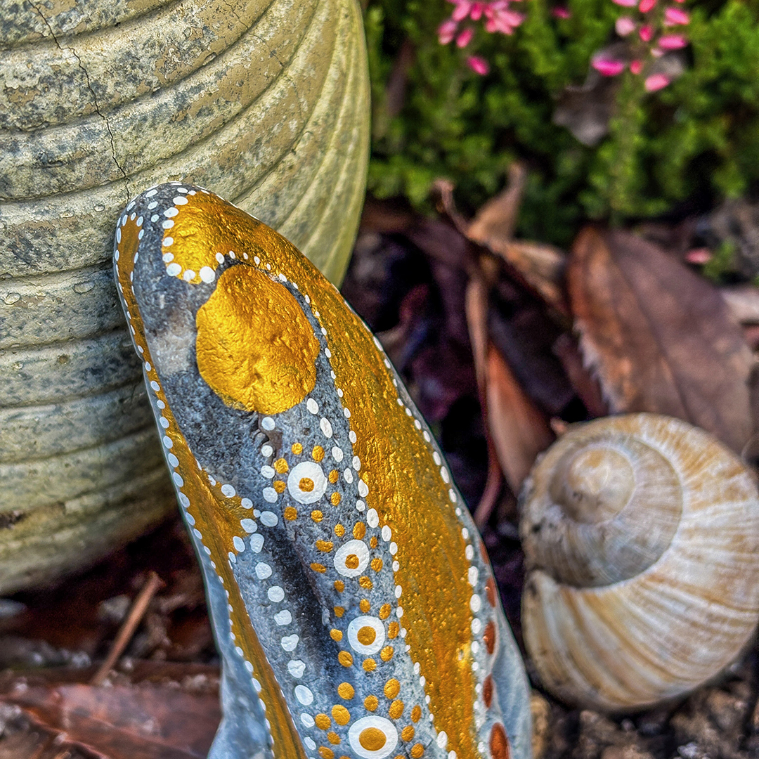 Bemalter Stein mit Engel vor roter Erikapflanze neben Schneckenhaus
