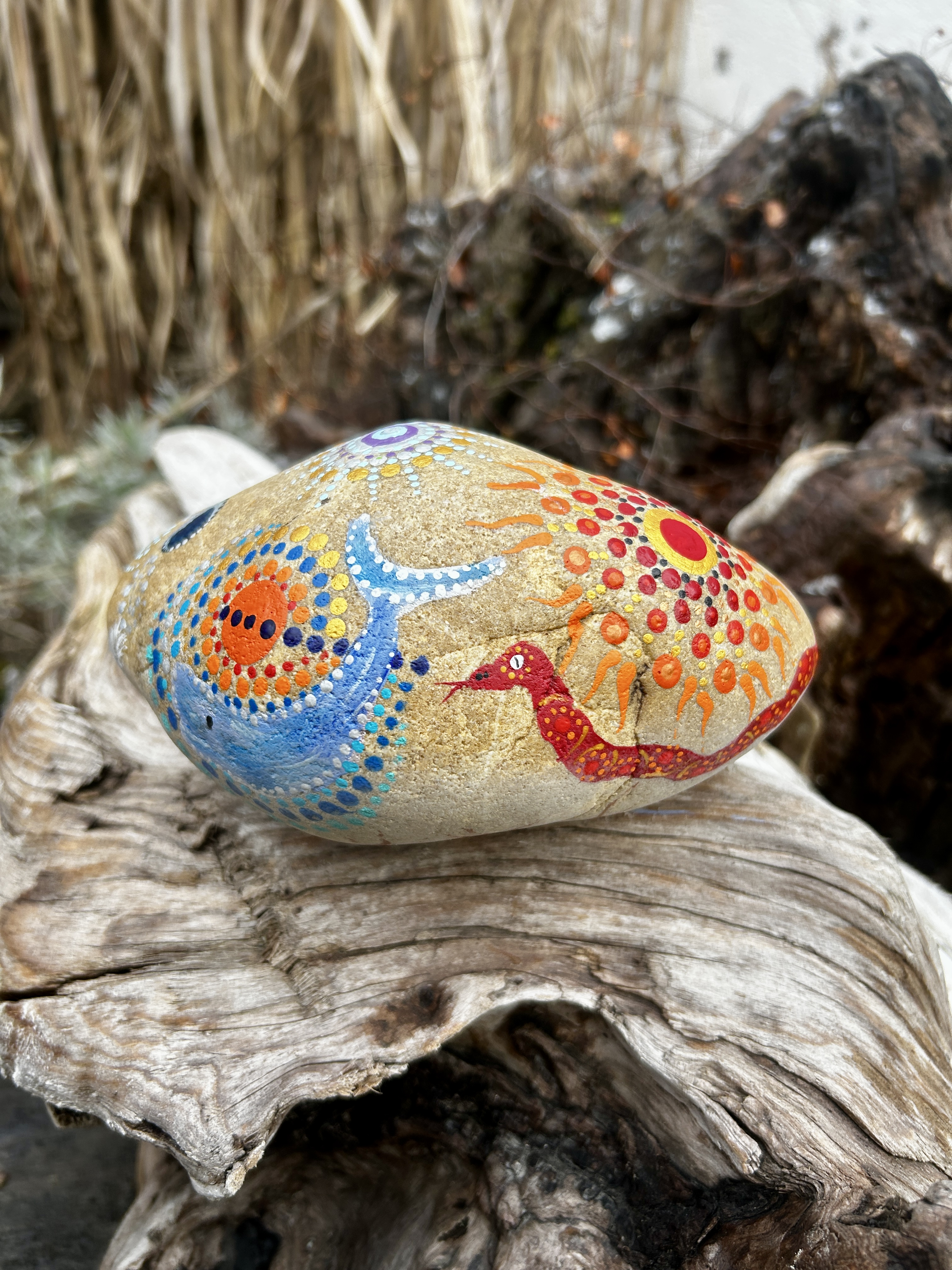 Stein in Dot Painting Technik bemalt mit Sonne, Schlange und Wal auf Wurzel liegend