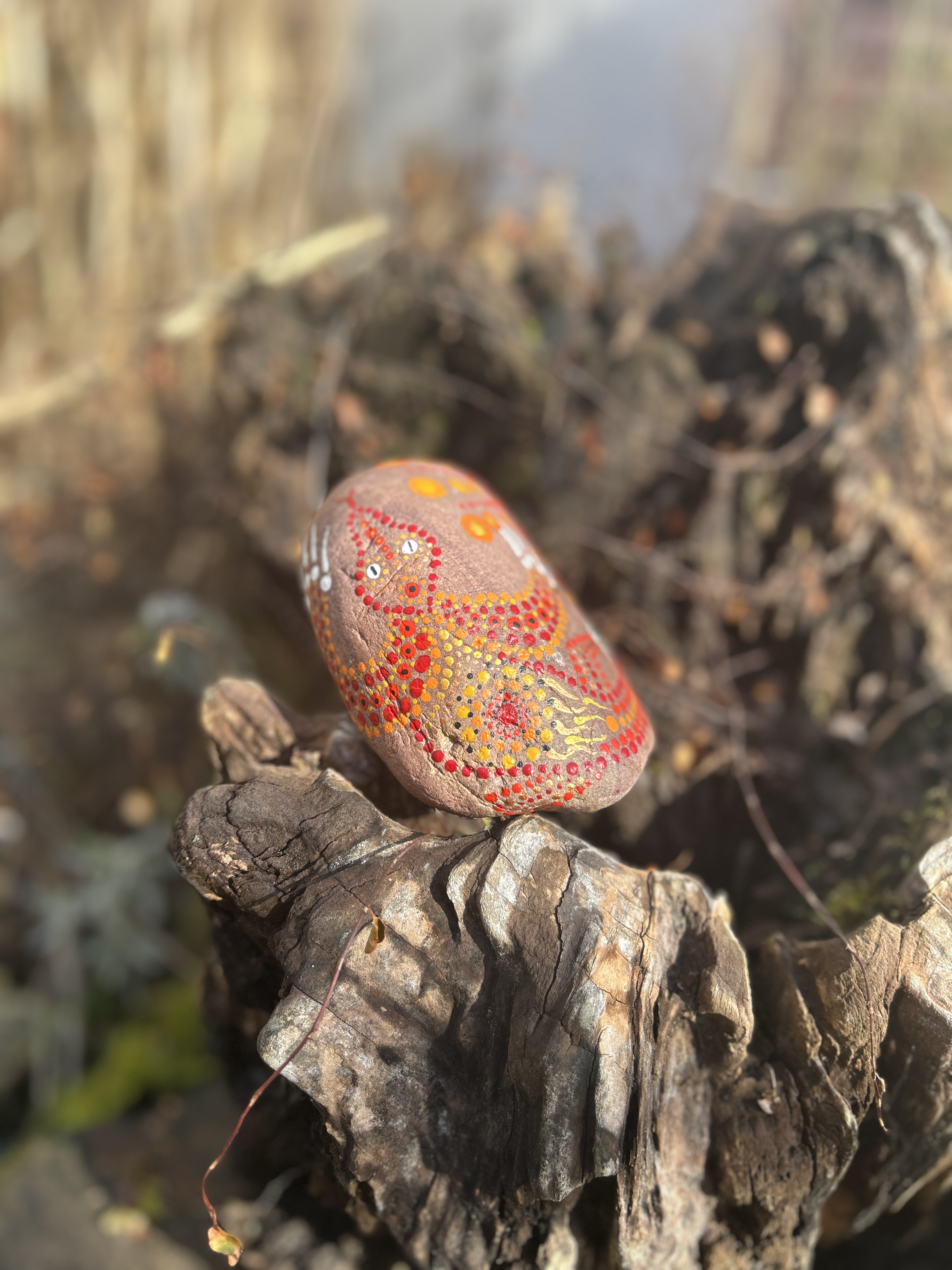 Rötlicher Stein in Dot Painting Techni  bemalt mit einem Drachen auf einer Wurzel liegend