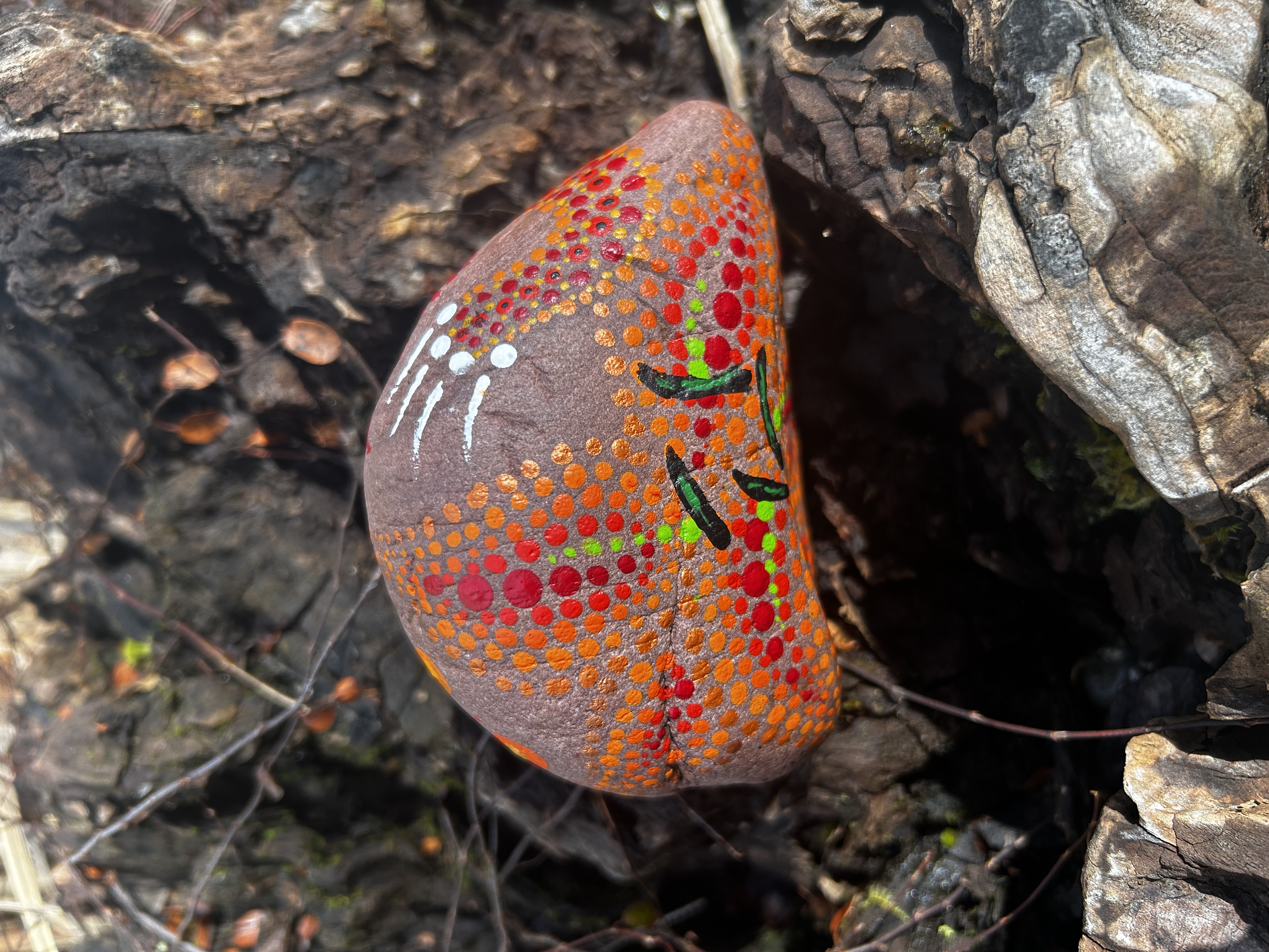  Rötlicher Stein in Dot Painting Technik  bemalt mit einer Blume auf einer Wurzel liegend