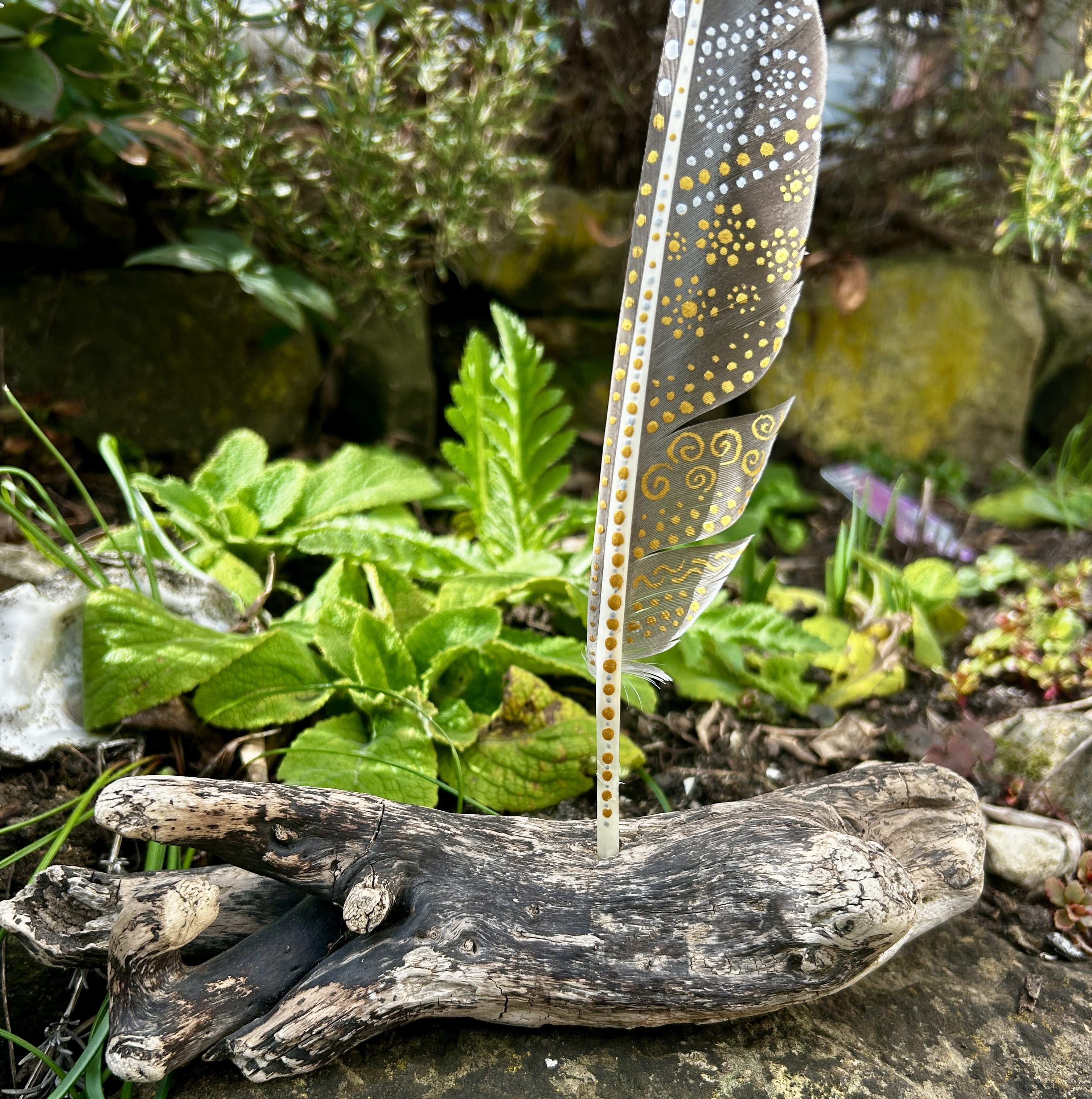 Eine bemalte Federn im Garten stehend in Silber und Gold gestaltet mit Symbolen in einem Ständer aus Treibholz