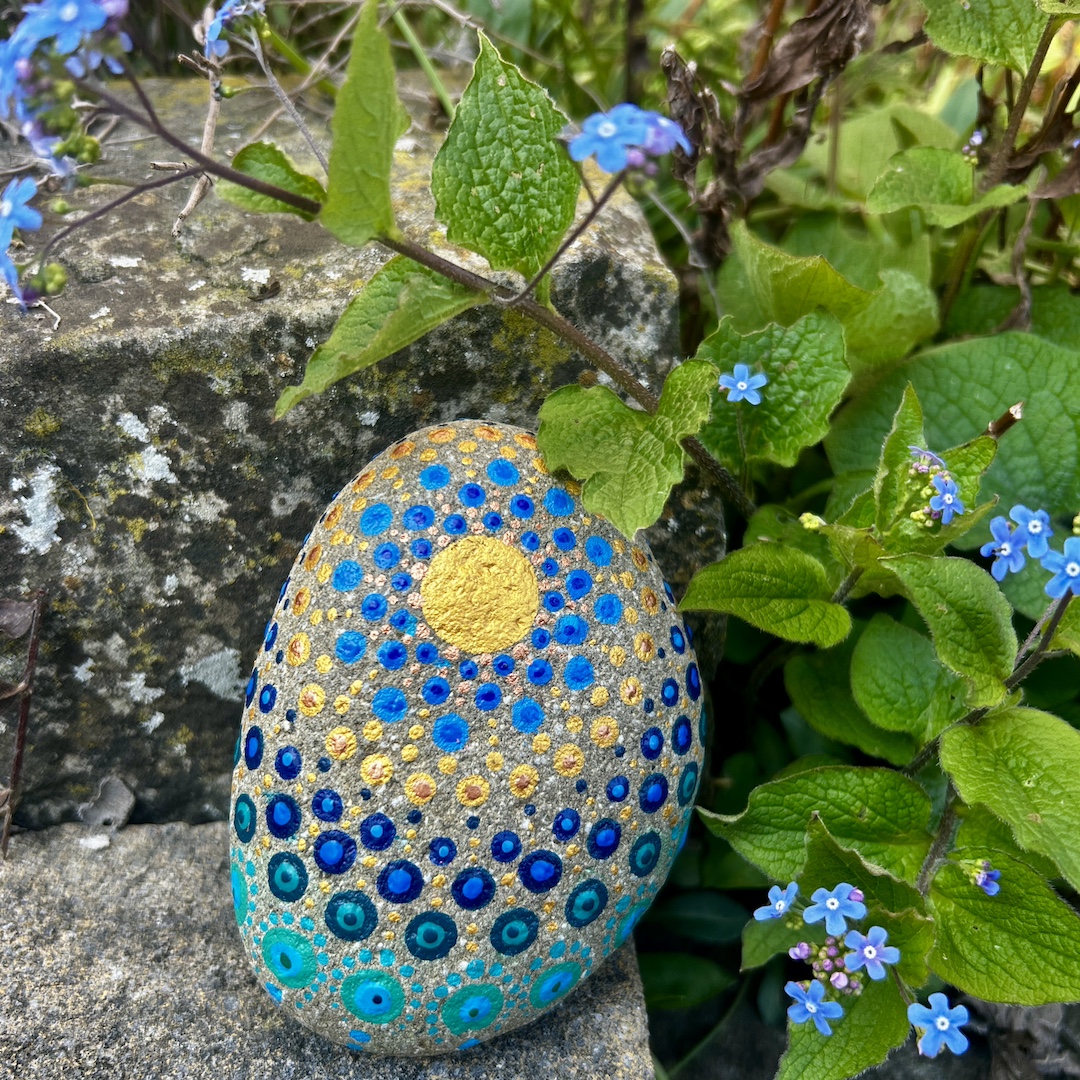 In Dot painting Technik bemalter Stein mit einem Mandala in Gold und Blautönen vor Vergissmeinnichts