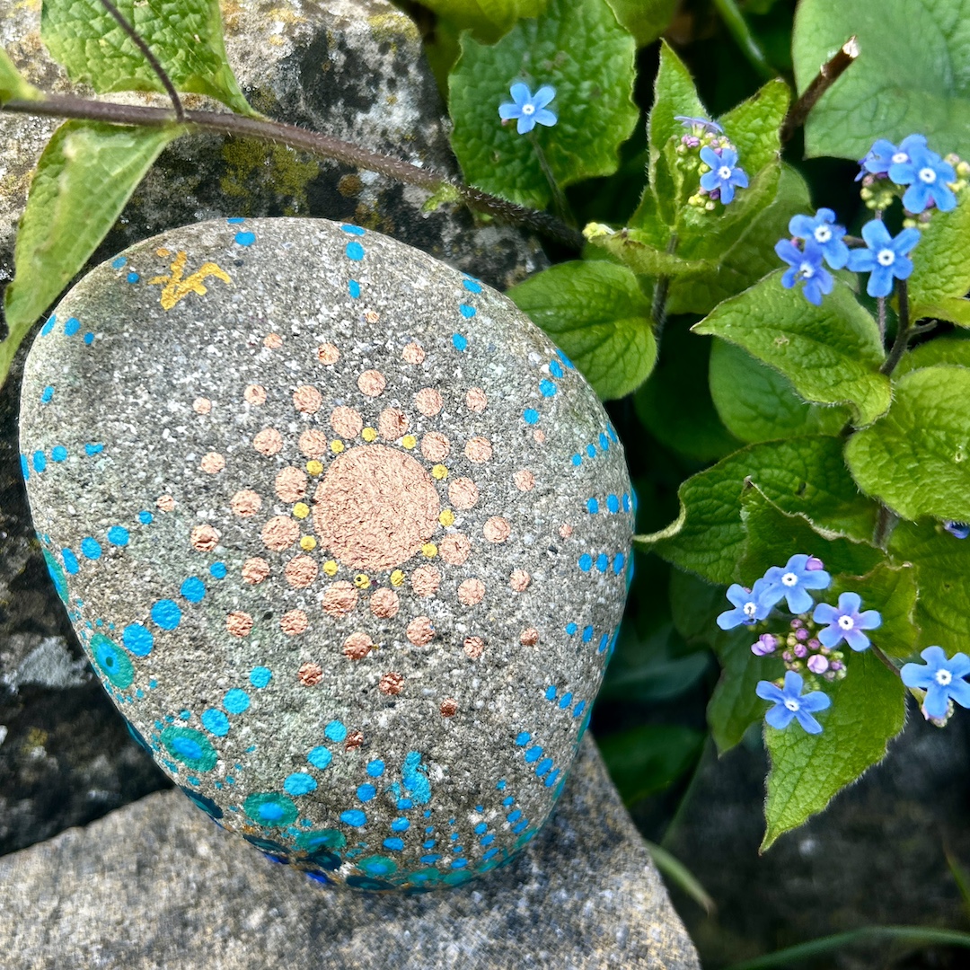 In Dot painting Technik bemalter Stein mit einem Mandala in Gold und Blautönen vor Vergissmeinnichts