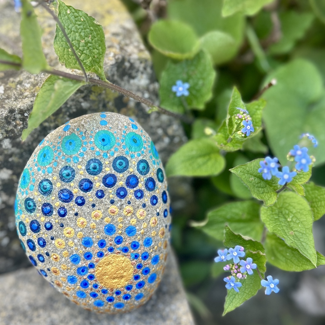 In Dot painting Technik bemalter Stein mit einem Mandala in Gold und Blautönen vor Vergissmeinnichts
