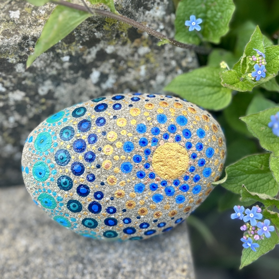 In Dot painting Technik bemalter Stein mit einem Mandala in Gold und Blautönen vor Vergissmeinnichts