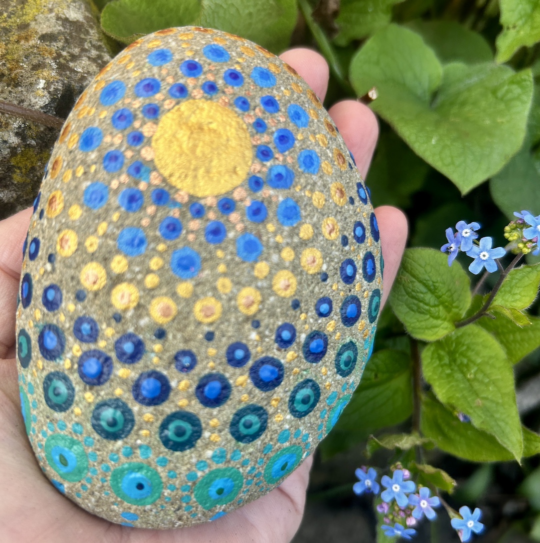 In Dot painting Technik bemalter Stein mit einem Mandala in Gold und Blautönen in der Hand gehalten. im Hintergrund Vergissmeinnichts.