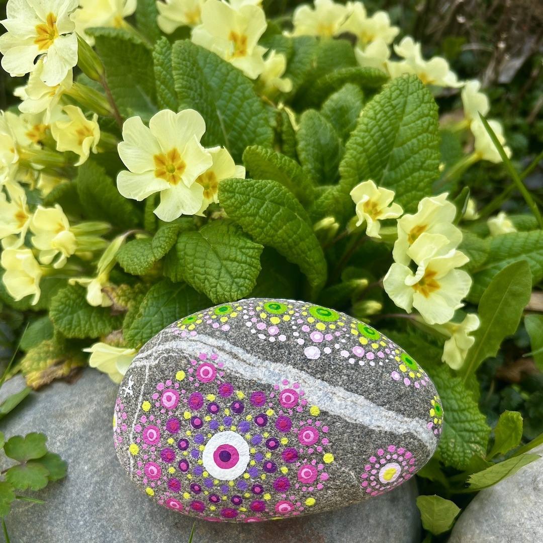 In Dot painting Technik bemalter Stein mit einem Mandala in Rosa-, Grün- und Gelbtönen vor Primeln