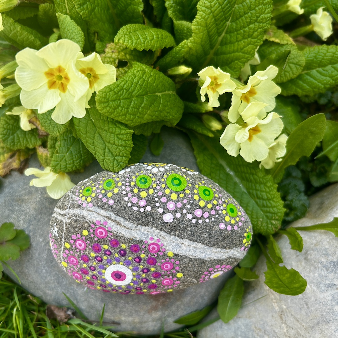 In Dot painting Technik bemalter Stein mit einem Mandala in Rosa-, Grün- und Gelbtönen vor Primeln