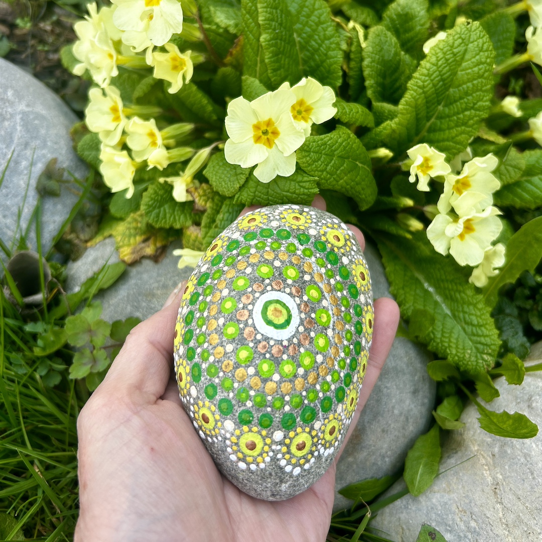 In Dot painting Technik bemalter Stein mit einem Mandala in Grün- und Gelbtönen in der Hand gehalten und im Hintergrund Primeln