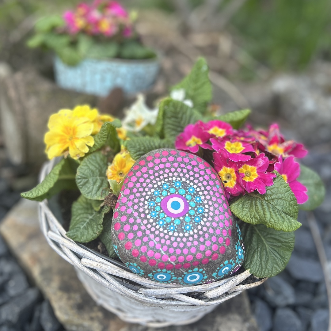 In Dot painting Technik bemalter Stein mit einem Mandala in Rosa- und Türkistönen in einem Körbchen liegend vor Primeln
