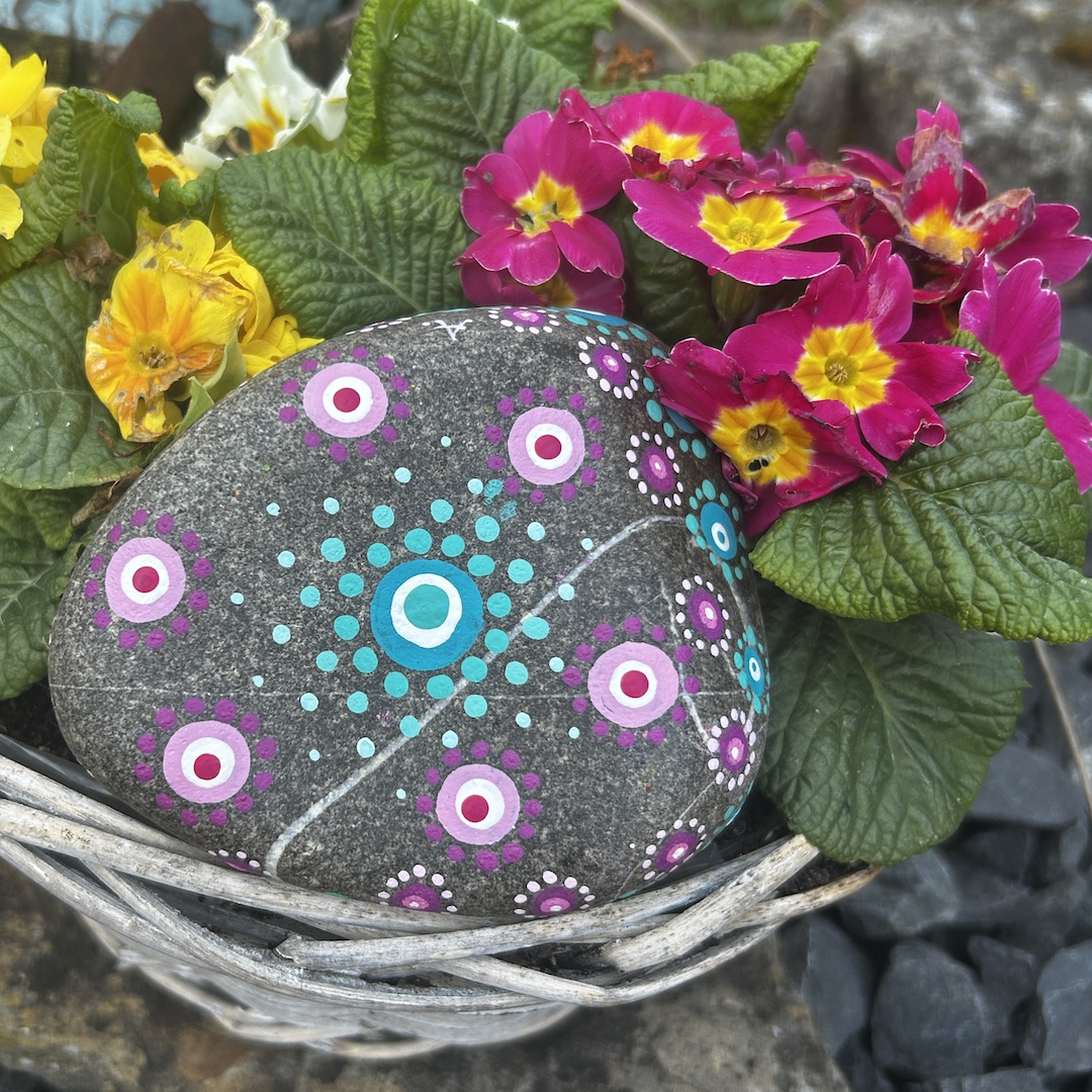 In Dot painting Technik bemalter Stein mit einem Mandala in Rosa- und Türkistönen vor Primeln