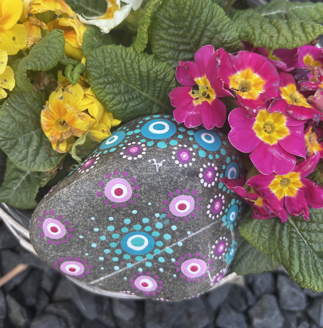 In Dot painting Technik bemalter Stein mit einem Mandala in Rosa- und Türkistönen vor Primeln