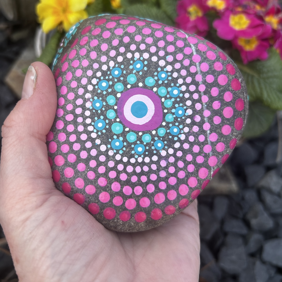 In Dot painting Technik bemalter Stein mit einem Mandala in Rosa- und Türkistönen in der Hand gehalten vor Primeln
