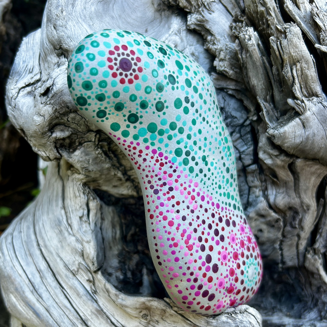 Stein mit einem mintfarbenen und magentafarbenen Mandala in Dot Painting technik bemalt an einer Wurzel lehnend.