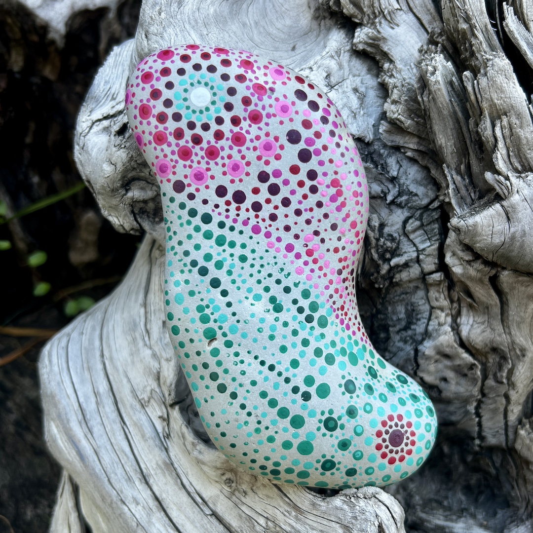 Stein mit einem mintfarbenen und magentafarbenen Mandala in Dot Painting technik bemalt an einer Wurzel lehnend.