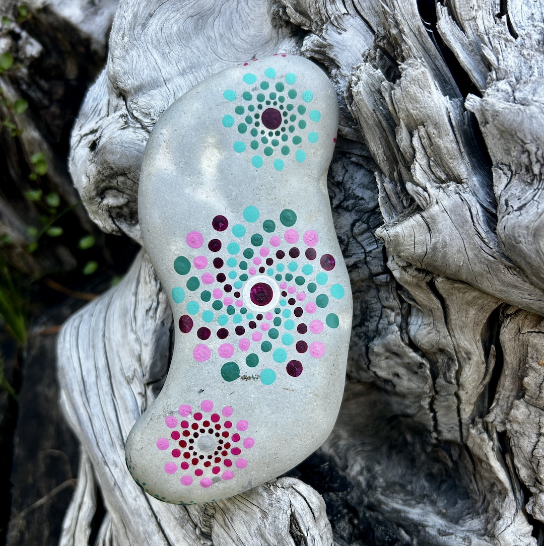 Stein mit einem mintfarbenen und magentafarbenen Mandala in Dot Painting technik bemalt an einer Wurzel lehnend.