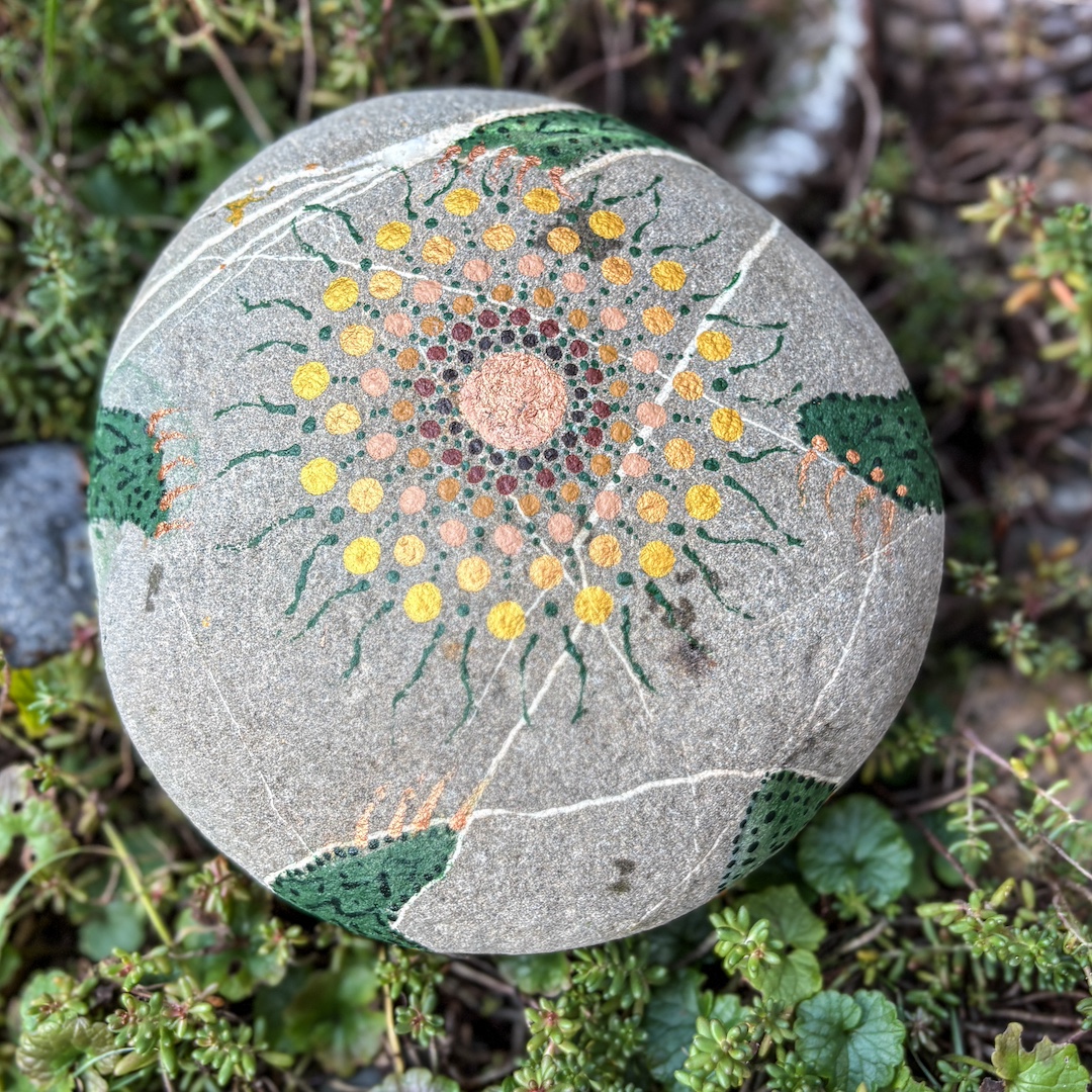 Stein mit Mandala in Erdfarben bemalt auf Grünpflanzen liegend