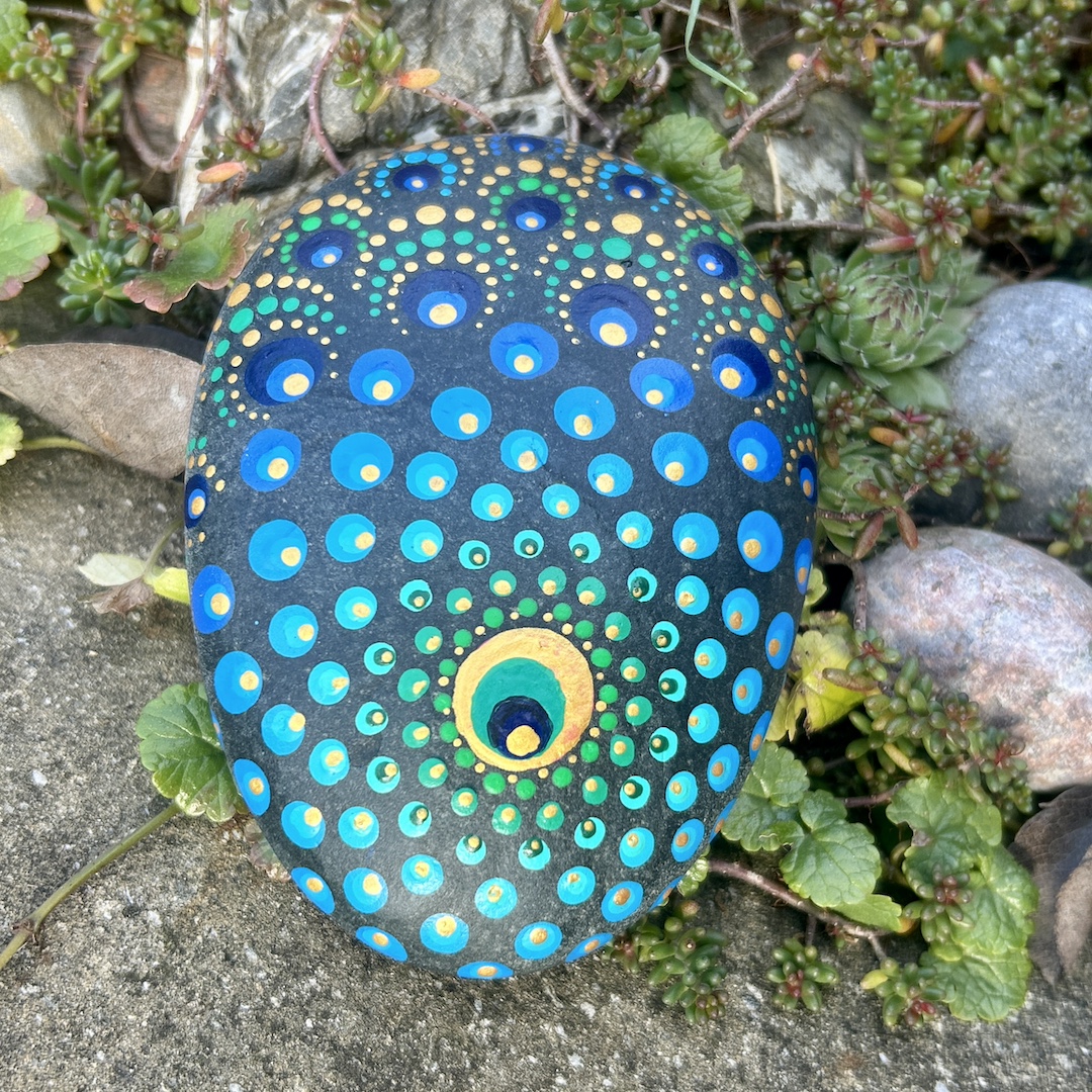 Stein mit einem Mandala bemalt in Farbtönen einer Pfauenfeder im Garten liegend