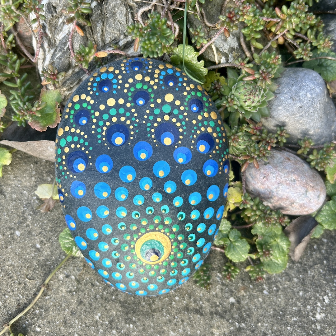 Stein mit einem Mandala bemalt in Farbtönen einer Pfauenfeder im Garten liegend
